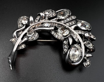 May include: A silver-toned brooch in the shape of a stylized leaf with clear rhinestones. The brooch is set with several large, faceted rhinestones and smaller rhinestones.