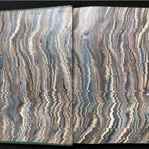 May include: Open book with marbled paper pages. The pages display a swirling pattern of blues, grays, and whites, creating a textured, wave-like effect. The book's spine is a muted green.