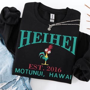May include: Black sweatshirt with the word "HEIHEI" in teal lettering. Below the text is a cartoon rooster and the text "EST. 2016 MOTUNUI, HAWAII" in pink and teal. A silver necklace is draped across the sweatshirt.