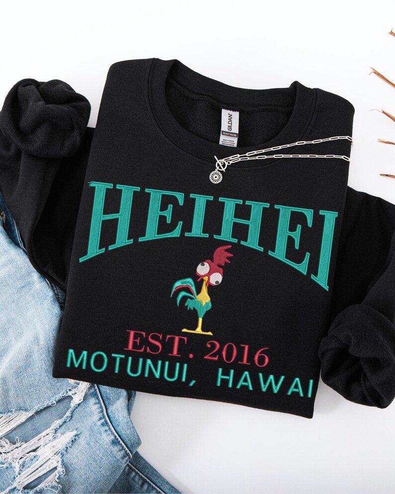 May include: Black sweatshirt with the word "HEIHEI" in teal lettering. Below the text is a cartoon rooster and the text "EST. 2016 MOTUNUI, HAWAII" in pink and teal. A silver necklace is draped across the sweatshirt.