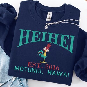 May include: Navy blue sweatshirt with the word "HEIHEI" in green arched lettering. Below is a cartoon rooster and the text "EST. 2016 MOTUNUI, HAWAII" in red and green. A silver necklace rests on the sweatshirt.