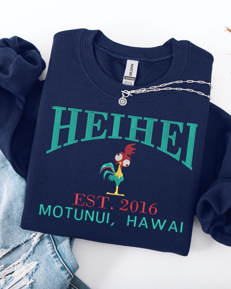May include: Navy blue sweatshirt with the word "HEIHEI" in green arched lettering. Below is a cartoon rooster and the text "EST. 2016 MOTUNUI, HAWAII" in red and green. A silver necklace rests on the sweatshirt.