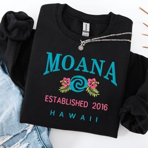 K&ouml;nnte beinhalten: Schwarzes Sweatshirt mit Rundhalsausschnitt, dem Wort "MOANA" in T&uuml;rkis, einem t&uuml;rkisfarbenen und wei&szlig;en Wirbeldesign, rosa Hibiskusbl&uuml;ten und dem Text "ESTABLISHED 2016 HAWAII". Eine silberne Halskette liegt auf dem Sweatshirt.