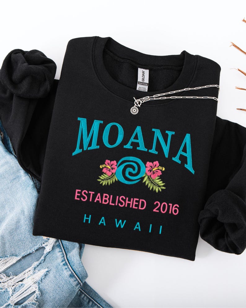 K&ouml;nnte beinhalten: Schwarzes Sweatshirt mit Rundhalsausschnitt, dem Wort "MOANA" in T&uuml;rkis, einem t&uuml;rkisfarbenen und wei&szlig;en Wirbeldesign, rosa Hibiskusbl&uuml;ten und dem Text "ESTABLISHED 2016 HAWAII". Eine silberne Halskette liegt auf dem Sweatshirt.