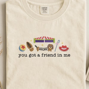 May include: Cream-colored t-shirt with embroidered cartoon images and the phrase "you got a friend in me." The design includes a ball, a cowboy boot, a slinky, a dog, a hat, and other toy-themed elements.