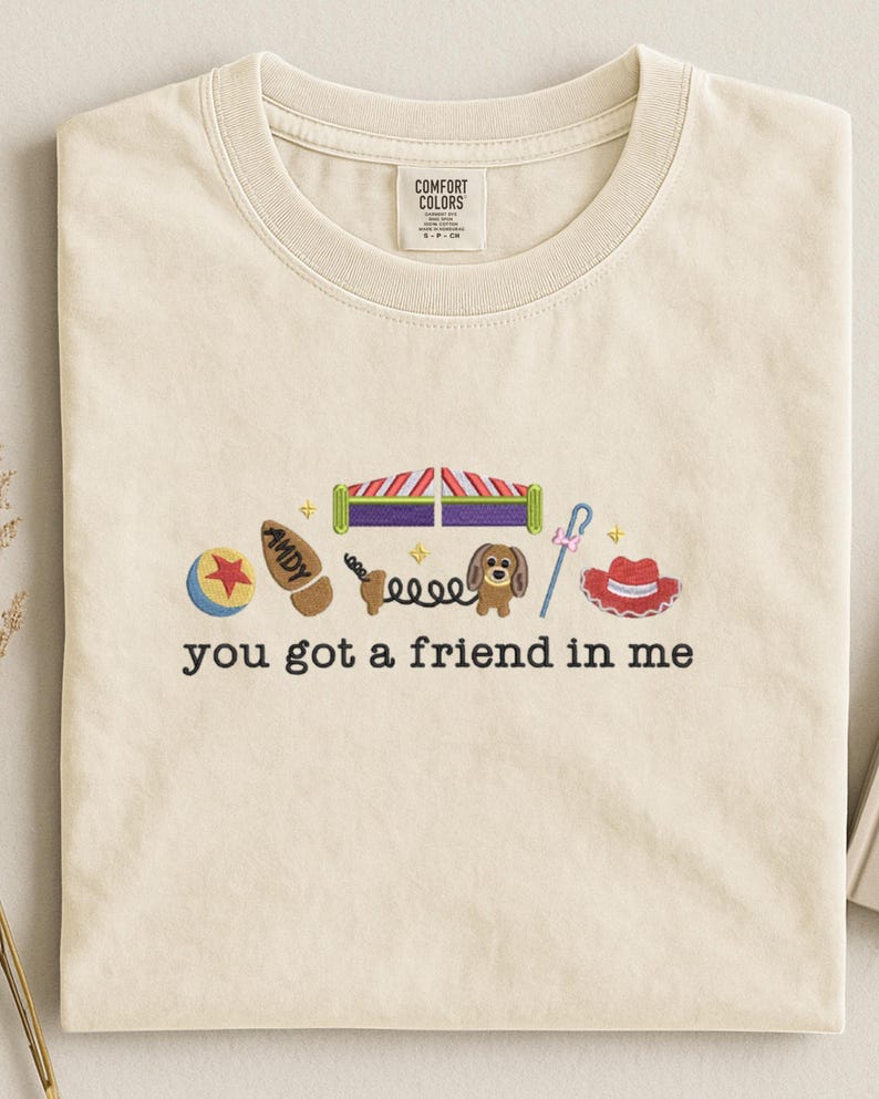 May include: Cream-colored t-shirt with embroidered cartoon images and the phrase "you got a friend in me." The design includes a ball, a cowboy boot, a slinky, a dog, a hat, and other toy-themed elements.