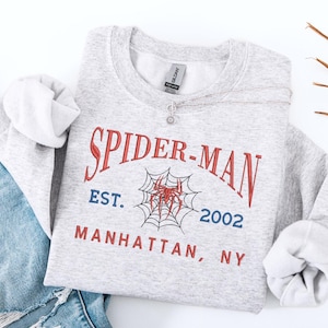 Może przedstawiać: Jasnoszara bluza Spider-Man z czerwonym i niebieskim tekstem oraz grafiką pajęczyny. Tekst brzmi "SPIDER-MAN EST. 2002 MANHATTAN, NY." Srebrny naszyjnik z wisiorkiem w kształcie kwiatu znajduje się na bluzie. Widoczne są niebieskie dżinsy.