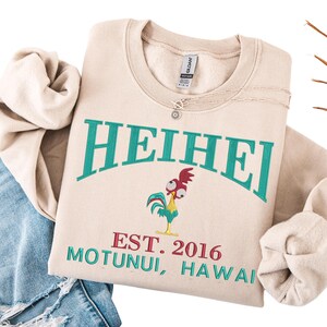 May include: Beige crewneck sweatshirt with the word "HEIHEI" in teal lettering. Below the word is a cartoon rooster and the text "EST. 2016 MOTUNUI, HAWAII" in red and teal. A silver necklace is on the sweatshirt.