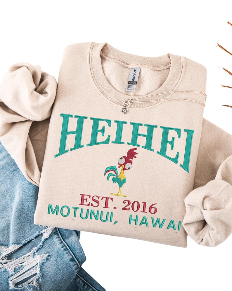 May include: Beige crewneck sweatshirt with the word "HEIHEI" in teal lettering. Below the word is a cartoon rooster and the text "EST. 2016 MOTUNUI, HAWAII" in red and teal. A silver necklace is on the sweatshirt.