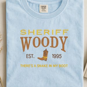 Puede incluir: Camiseta azul claro con la inscripci&oacute;n bordada "SHERIFF WOODY" en amarillo y marr&oacute;n. Debajo, "EST. 1995" y una bota de vaquero. La frase "THERE'S A SNAKE IN MY BOOT" est&aacute; bordada debajo de la bota.