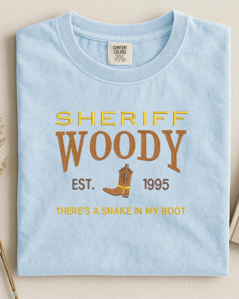 Puede incluir: Camiseta azul claro con la inscripci&oacute;n bordada "SHERIFF WOODY" en amarillo y marr&oacute;n. Debajo, "EST. 1995" y una bota de vaquero. La frase "THERE'S A SNAKE IN MY BOOT" est&aacute; bordada debajo de la bota.