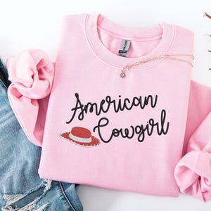 May include: A pale pink sweatshirt featuring the text "American Cowgirl" in black script, with a red and white cowboy hat graphic. A gold necklace with a small pendant is also visible.