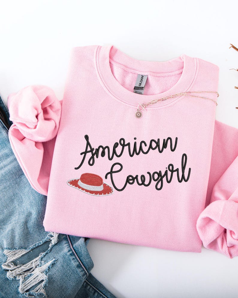 May include: A pale pink sweatshirt featuring the text "American Cowgirl" in black script, with a red and white cowboy hat graphic. A gold necklace with a small pendant is also visible.