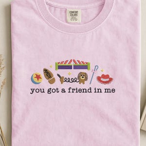 May include: Light pink t-shirt with embroidered images of a ball, boot, toy box, dog, and hat. The text "you got a friend in me" is embroidered below the images. The shirt has a crew neck and short sleeves.