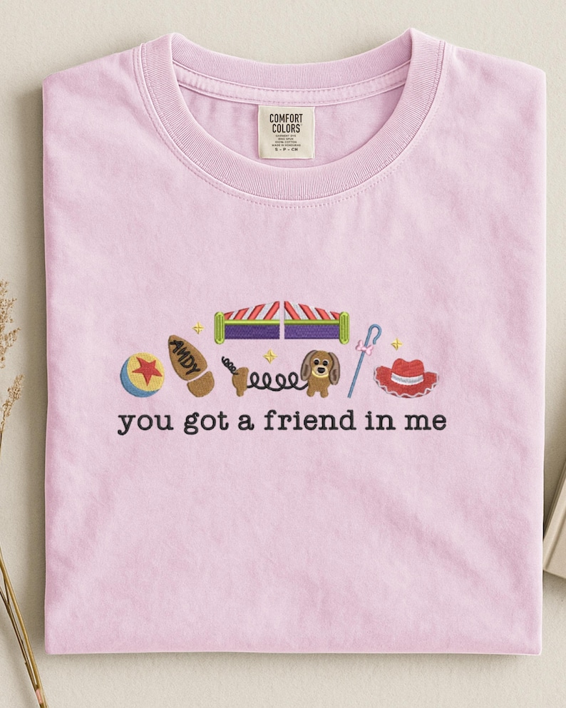 May include: Light pink t-shirt with embroidered images of a ball, boot, toy box, dog, and hat. The text "you got a friend in me" is embroidered below the images. The shirt has a crew neck and short sleeves.