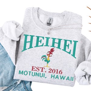 May include: A light gray sweatshirt with the word "HEIHEI" in teal arched lettering. Below the text is a cartoon rooster and the text "EST. 2016 MOTUNUI, HAWAII" in red and teal. A silver necklace with a small pendant is on the sweatshirt.