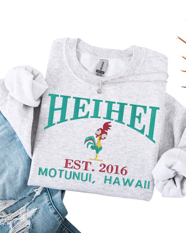 May include: A light gray sweatshirt with the word "HEIHEI" in teal arched lettering. Below the text is a cartoon rooster and the text "EST. 2016 MOTUNUI, HAWAII" in red and teal. A silver necklace with a small pendant is on the sweatshirt.