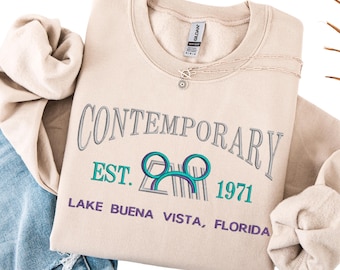 May include: Beige sweatshirt with the word "CONTEMPORARY" embroidered in gray, along with "EST. 1971" and "LAKE BUENA VISTA, FLORIDA" in teal and purple. A silver necklace with a sun pendant is on the sweatshirt.