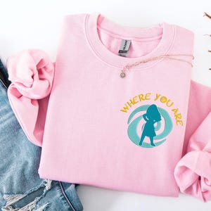 Może przedstawiać: Jasnor&oacute;żowa bluza z napisem "WHERE YOU ARE" i turkusowo-ż&oacute;łtą grafiką. Bluza jest złożona z parą niebieskich dżins&oacute;w. Naszyjnik z małą zawieszką leży na bluzie.