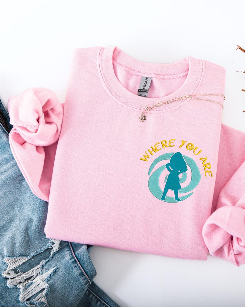 Może przedstawiać: Jasnor&oacute;żowa bluza z napisem "WHERE YOU ARE" i turkusowo-ż&oacute;łtą grafiką. Bluza jest złożona z parą niebieskich dżins&oacute;w. Naszyjnik z małą zawieszką leży na bluzie.