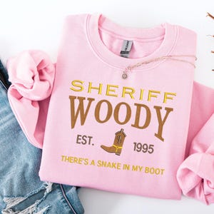 Puede incluir: Sudadera rosa claro con el texto "SHERIFF WOODY" en letras doradas y marrones, con un dibujo de bota de vaquero. Tambi&eacute;n se muestra el texto "EST. 1995" y "THERE'S A SNAKE IN MY BOOT". Un collar con un peque&ntilde;o colgante est&aacute; sobre la sudadera.