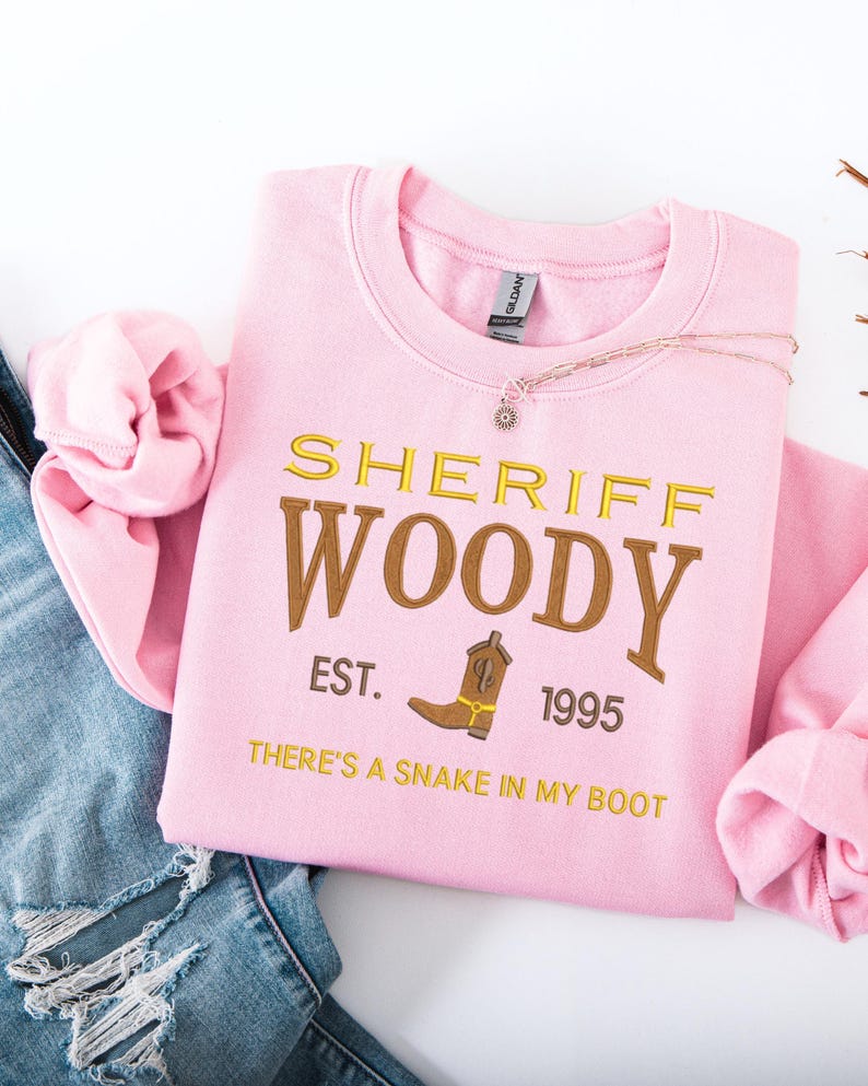 Puede incluir: Sudadera rosa claro con el texto "SHERIFF WOODY" en letras doradas y marrones, con un dibujo de bota de vaquero. Tambi&eacute;n se muestra el texto "EST. 1995" y "THERE'S A SNAKE IN MY BOOT". Un collar con un peque&ntilde;o colgante est&aacute; sobre la sudadera.