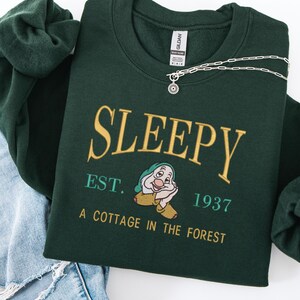 Peut inclure: Un sweat-shirt vert foncé avec le mot "SLEEPY" en lettres dorées, ainsi que "EST. 1937" et "A COTTAGE IN THE FOREST". Le sweat-shirt présente un dessin de personnage de dessin animé et est accompagné d'un collier argenté.