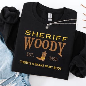 Puede incluir: Sudadera negra con el texto "SHERIFF WOODY" en amarillo y marr&oacute;n, con un dibujo de bota de vaquero. Tambi&eacute;n se ve el texto "EST. 1995" y "THERE'S A SNAKE IN MY BOOT". Una cadena plateada est&aacute; sobre la sudadera.