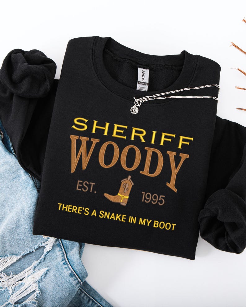 Puede incluir: Sudadera negra con el texto "SHERIFF WOODY" en amarillo y marr&oacute;n, con un dibujo de bota de vaquero. Tambi&eacute;n se ve el texto "EST. 1995" y "THERE'S A SNAKE IN MY BOOT". Una cadena plateada est&aacute; sobre la sudadera.