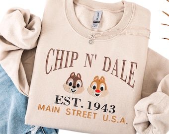 Puede incluir: Sudadera beige con bordados de los personajes Chip y Chop y el texto "Chip N' Dale EST. 1943 Main Street U.S.A." Un collar plateado con un colgante de sol está sobre la sudadera. La sudadera está doblada sobre unos vaqueros.