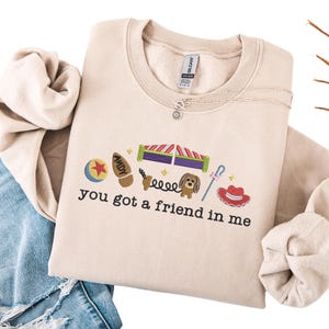 May include: Beige crewneck sweatshirt with embroidered cartoon characters and the phrase "you got a friend in me." The design includes a ball, a boot, a dog, and a cowboy hat. A silver necklace is visible.