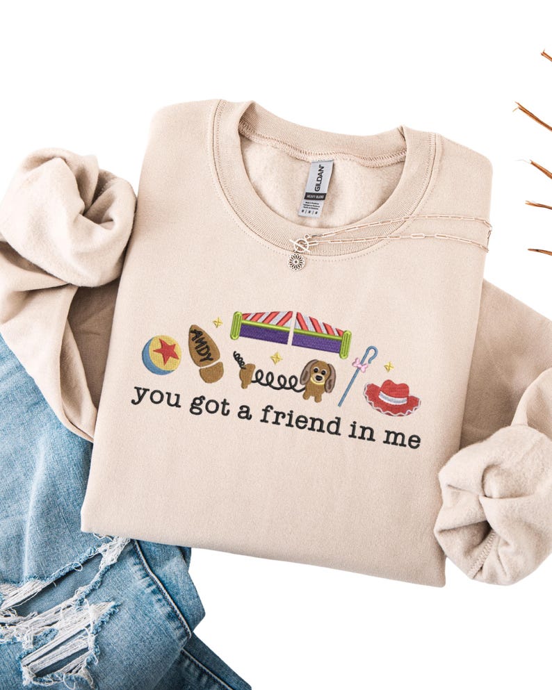 May include: Beige crewneck sweatshirt with embroidered cartoon characters and the phrase "you got a friend in me." The design includes a ball, a boot, a dog, and a cowboy hat. A silver necklace is visible.