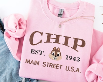Puede incluir: Sudadera rosa claro con la palabra "CHIP" bordada en marrón. Debajo, el texto "EST. 1943 MAIN STREET U.S.A." y un personaje de Chip bordado. Un collar dorado con un pequeño colgante descansa sobre la sudadera.
