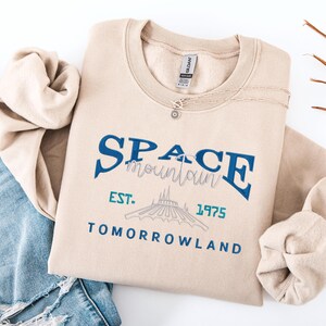 以下が含まれることがあります： ベージュ色のクルーネックのスウェットシャツ。青とグレーの文字で「SPACE mountain」と書かれています。その下に、青とティールで「EST. 1975 TOMORROWLAND」と書かれています。太陽のペンダントが付いたシルバーのネックレスがスウェットシャツの上にあります。スウェットシャツは青いジーンズの上に折りたたまれています。