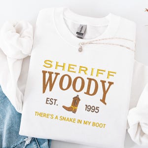Puede incluir: Sudadera blanca con el texto bordado "SHERIFF WOODY" en oro y marr&oacute;n, un gr&aacute;fico de bota de vaquero y las frases "EST. 1995" y "THERE'S A SNAKE IN MY BOOT". Un collar plateado est&aacute; drapeado sobre la sudadera.