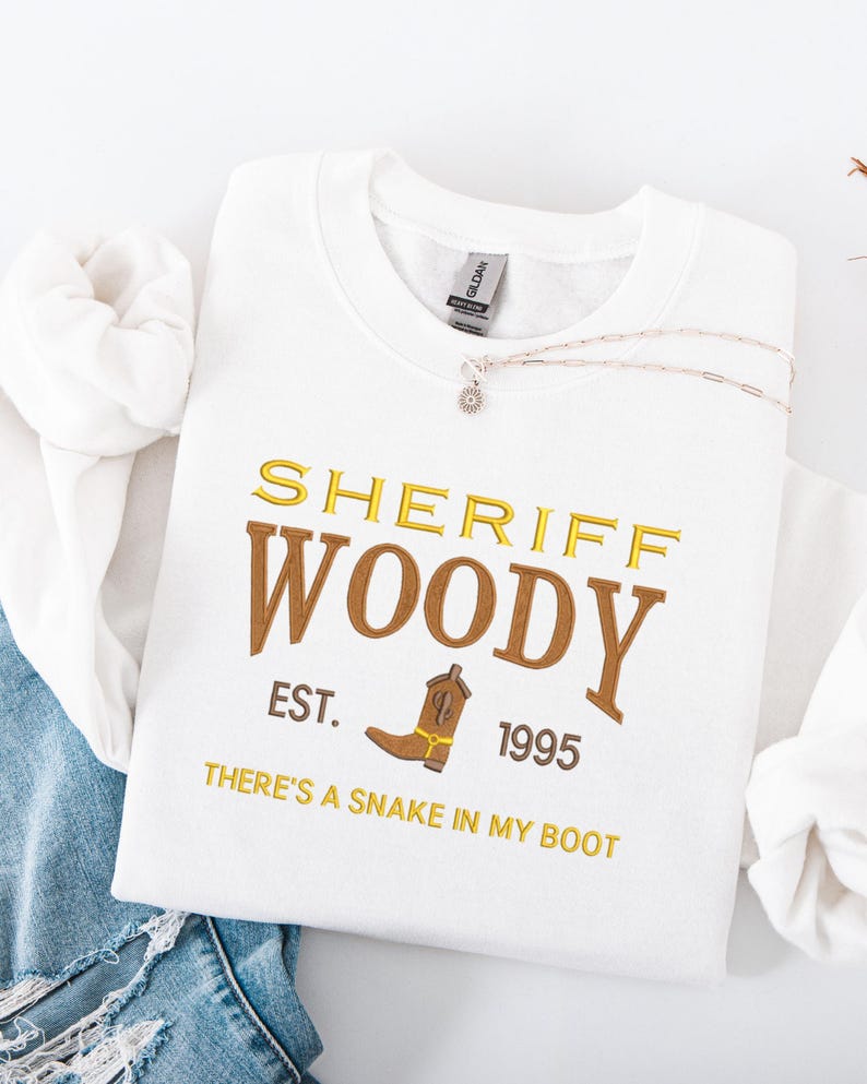 Puede incluir: Sudadera blanca con el texto bordado "SHERIFF WOODY" en oro y marr&oacute;n, un gr&aacute;fico de bota de vaquero y las frases "EST. 1995" y "THERE'S A SNAKE IN MY BOOT". Un collar plateado est&aacute; drapeado sobre la sudadera.