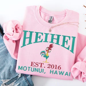 May include: A light pink sweatshirt with the word "HEIHEI" in teal arched across the top. Below the word is a cartoon rooster and the text "EST. 2016 MOTUNUI, HAWAII" in teal and red. A gold necklace with a sun pendant rests on the sweatshirt.