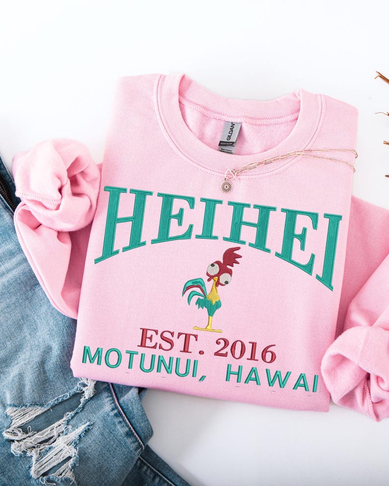 May include: A light pink sweatshirt with the word "HEIHEI" in teal arched across the top. Below the word is a cartoon rooster and the text "EST. 2016 MOTUNUI, HAWAII" in teal and red. A gold necklace with a sun pendant rests on the sweatshirt.