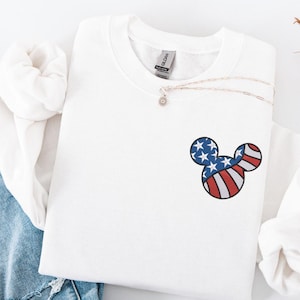 Peut inclure: Sweat-shirt blanc avec un petit motif t&ecirc;te de Mickey Mouse. Le motif reprend les couleurs du drapeau am&eacute;ricain : rayures rouges et blanches et bleu avec des &eacute;toiles blanches. Un collier argent&eacute; avec un petit pendentif est &eacute;galement visible.