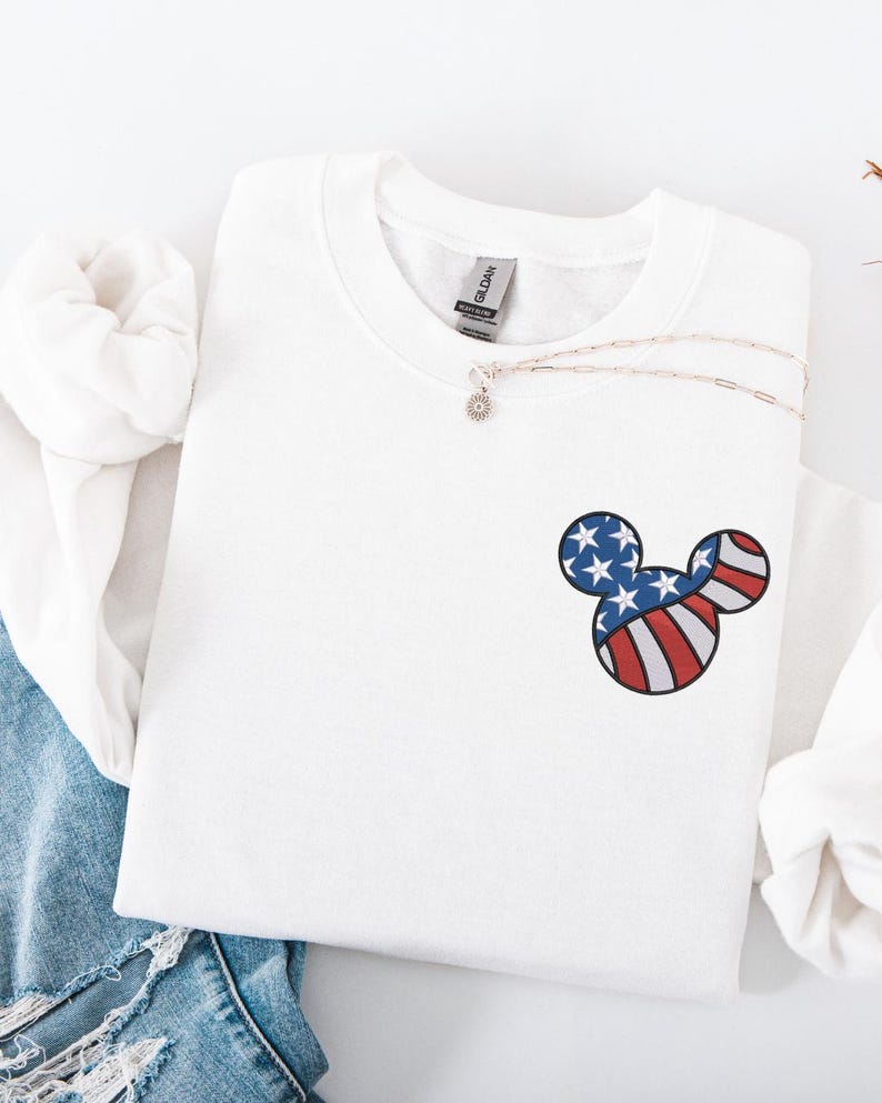 Peut inclure: Sweat-shirt blanc avec un petit motif t&ecirc;te de Mickey Mouse. Le motif reprend les couleurs du drapeau am&eacute;ricain : rayures rouges et blanches et bleu avec des &eacute;toiles blanches. Un collier argent&eacute; avec un petit pendentif est &eacute;galement visible.