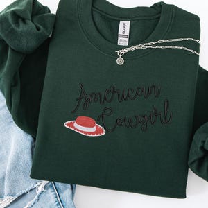 May include: A dark green sweatshirt featuring the embroidered phrase "American Cowgirl" in a script font, with a hat illustration. A silver necklace with a circular pendant is draped across the sweatshirt. The sweatshirt is folded on top of blue denim jeans.