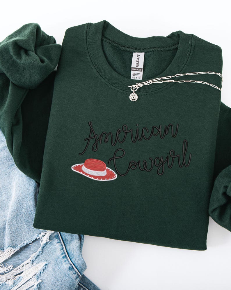 May include: A dark green sweatshirt featuring the embroidered phrase "American Cowgirl" in a script font, with a hat illustration. A silver necklace with a circular pendant is draped across the sweatshirt. The sweatshirt is folded on top of blue denim jeans.