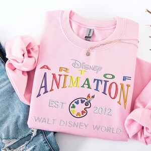 Pode incluir: Um moletom rosa claro com a inscrição bordada "Disney Art of Animation" em letras coloridas. O design inclui uma paleta de pintura e o texto "EST 2012 Walt Disney World". Um colar de ouro com um pequeno pingente repousa sobre o moletom.