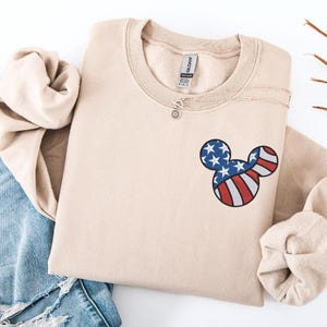 Peut inclure: Un sweat-shirt beige &agrave; col rond avec un motif de t&ecirc;te de Mickey Mouse rempli des couleurs du drapeau am&eacute;ricain. Un collier argent&eacute; avec un pendentif soleil est visible. Le sweat-shirt est pli&eacute;, et un jean bleu est en arri&egrave;re-plan.