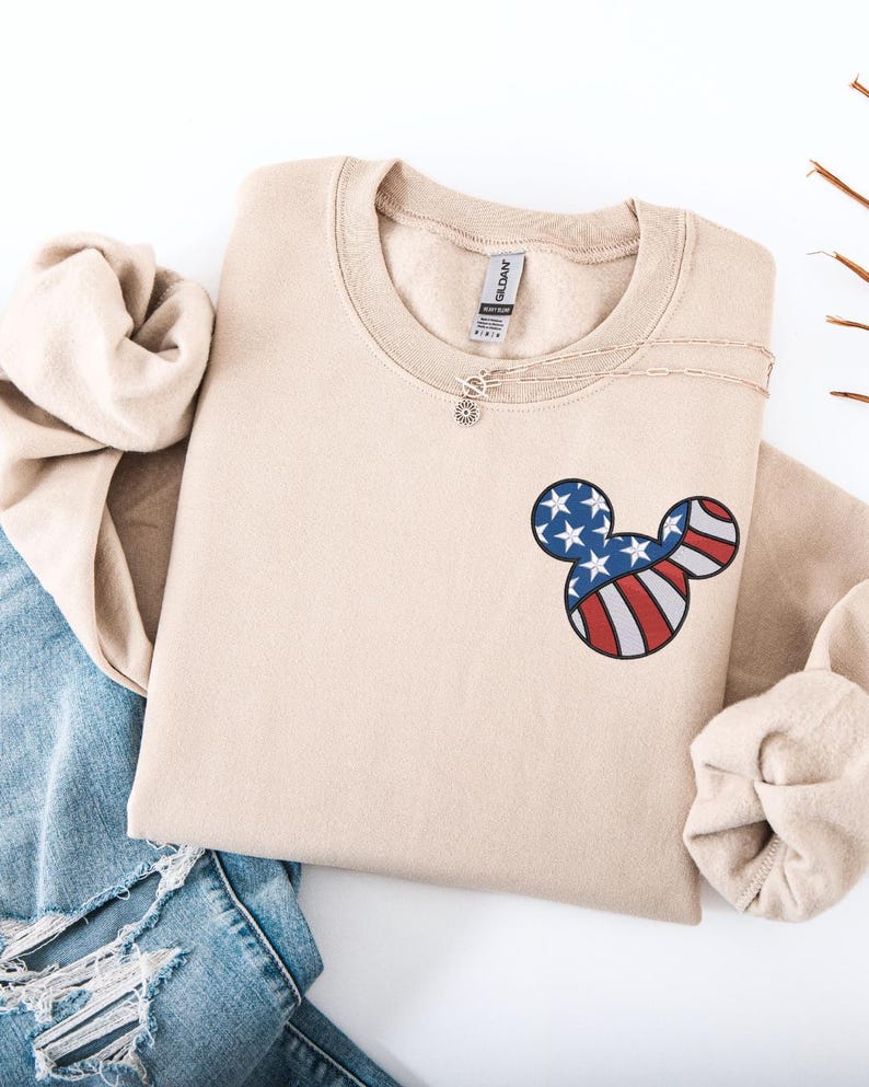 Peut inclure: Un sweat-shirt beige &agrave; col rond avec un motif de t&ecirc;te de Mickey Mouse rempli des couleurs du drapeau am&eacute;ricain. Un collier argent&eacute; avec un pendentif soleil est visible. Le sweat-shirt est pli&eacute;, et un jean bleu est en arri&egrave;re-plan.