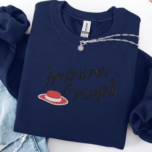 May include: A navy blue sweatshirt featuring the embroidered phrase "American Cowgirl" in a cursive font, with a red and white cowboy hat graphic. A silver necklace with a sun pendant is placed on the sweatshirt. The sweatshirt is folded on blue denim jeans.