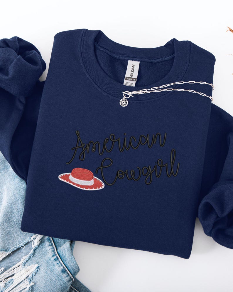 May include: A navy blue sweatshirt featuring the embroidered phrase "American Cowgirl" in a cursive font, with a red and white cowboy hat graphic. A silver necklace with a sun pendant is placed on the sweatshirt. The sweatshirt is folded on blue denim jeans.