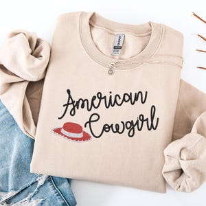May include: A beige sweatshirt featuring the text "American Cowgirl" in black script, and a red and white cowboy hat graphic. A delicate necklace is placed on the sweatshirt. The sweatshirt is folded on top of blue denim jeans.