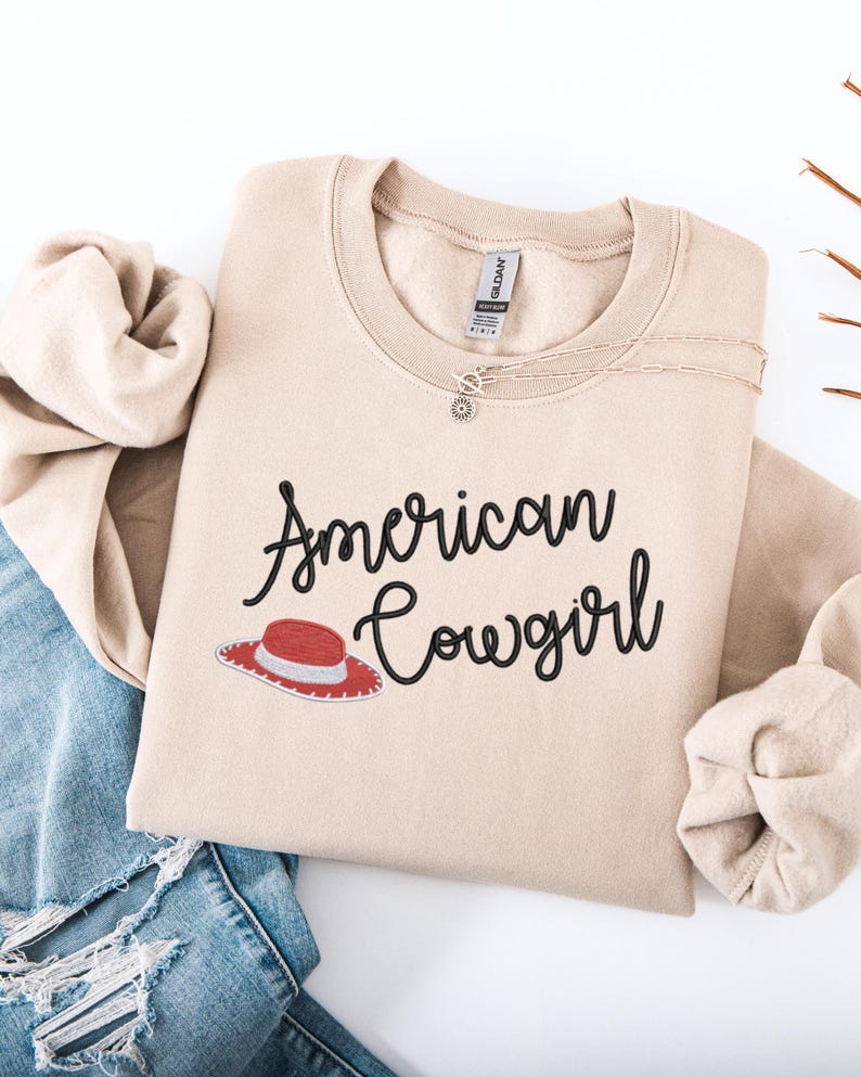 May include: A beige sweatshirt featuring the text "American Cowgirl" in black script, and a red and white cowboy hat graphic. A delicate necklace is placed on the sweatshirt. The sweatshirt is folded on top of blue denim jeans.