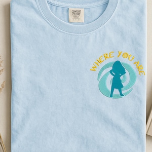 Może przedstawiać: Jasnoniebieska koszulka Comfort Colors z haftowanym napisem "WHERE YOU ARE" i turkusową sylwetką osoby. Koszulka ma okrągły dekolt i małą metkę na g&oacute;rnej lewej piersi.